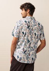 Camicia a maniche corte in tessuto leggero con motivo floreale e foglie multicolori su sfondo chiaro, con colletto classico e orlo arrotondato.