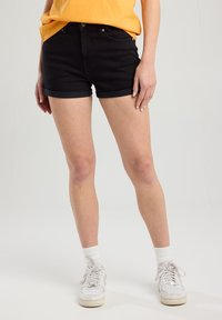 Shorts en denim noir avec ourlets retroussés, poches avant et quincaillerie argentée, associés à une chemise jaune et des baskets blanches.