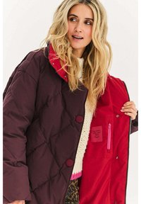 Fioletowa pikowana kurtka z czerwoną podszewką, dużymi guzikami i bocznymi kieszeniami. W środku znajduje się widoczna różowa kieszeń na zamek z metką.
