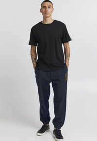 Mann med kort hår iført svart t-skjorte, marineblå joggebukser med bokstaven "S" og svarte joggesko, står med hendene i lommene.