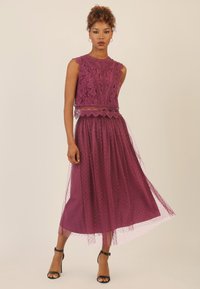 Paarse jurk met een kant bodice met een gegolfde rand, gelaagd met een tule rok met stippen. Zwarte sleehakken worden gedragen.