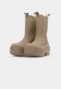 Botas de tobillo beige de material de ante suave, con paneles laterales elásticos, suelas de goma gruesas y un emblema circular en el lateral.