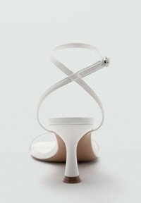 Białe sandały na obcasie z okrągłym noskiem, cienkimi paskami wokół kostki i stożkowym obcasem. Cholewka charakteryzuje się gładkim, błyszczącym wykończeniem z minimalną ilością metalowych elementów.