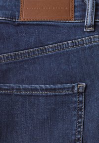 Jeans de mezclilla azul oscuro con un parche de cuero marrón que dice "STREET ONE STUDIO". Cuenta con costuras visibles y un bolsillo frontal.