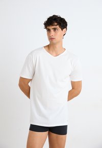 T-shirt en coton blanc à manches courtes et col en V, coupe décontractée, avec un détail de couture subtil au niveau du col, porté avec un short noir.