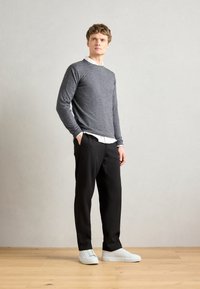 Suéter gris de manga larga sobre una camisa blanca con collar, combinado con pantalones negros y zapatillas deportivas blancas sobre un suelo de madera contra un fondo neutro.