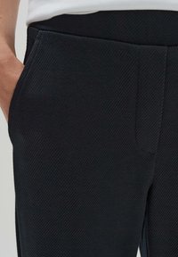 Svarta ribbstickade byxor med snedställda fickor och en slät midjeband, som visar ett texturerat tyg med ett subtilt diagonalt mönster.