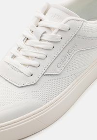 Sneaker bianco realizzato in mesh testurizzato e pelle, presenta lacci piatti, una marca discreta e una suola in gomma liscia. Design minimalista.