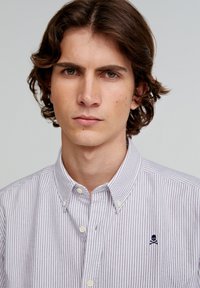 Cămașă cu dungi, cu butoni, de culoare mov deschis și alb. Are un guler ascuțit și un logo brodat cu craniu pe piept. Confort standard.
