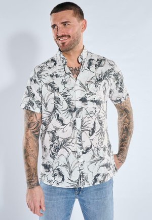 Man with short hair and tattoos wearing a white short-sleeve button-up shirt with a black floral pattern and blue jeans.