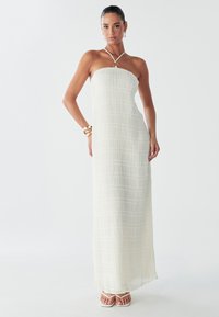 Witte strapless maxi-jurk met getextureerde verticale plooien, een halternek met een cirkelvormig accent, gecombineerd met witte sandalen.