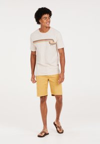 Camiseta color beige claro con un diseño de olas colorido, combinada con pantalones cortos amarillos. El modelo lleva chanclas marrones y está de pie frente a un fondo liso.