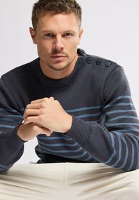 Maglione a maglia blu navy con righe orizzontali blu, polsini a coste e bottoni sulle spalle. Il modello è seduto con le mani intrecciate sopra un tessuto bianco.