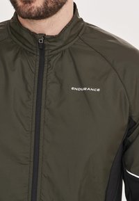 Olivgrön vindjacka med svarta accenter, med hög krage och dragkedja, och "ENDURANCE"-logotypen i vitt på bröstet.