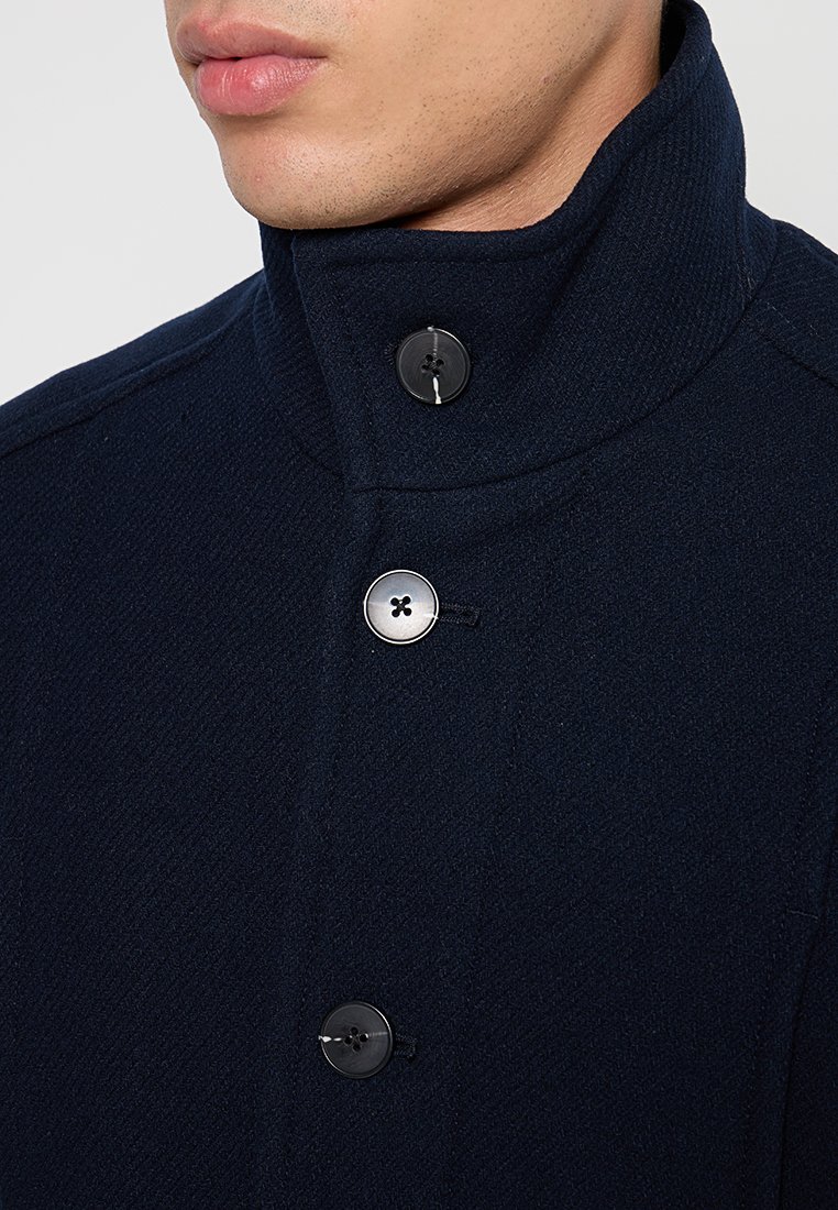 Marineblauwe wollen jas met hoge kraag, voorzien van zwarte knopen en een gestructureerde afwerking. Close-up weergave die knopen en het ontwerp van de kraag benadrukt.