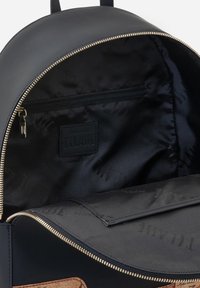 Interno dello zaino nero con una tasca con zip, rivestimento liscio e etichetta del marchio. Dettaglio della zip in ottone e base marrone a contrasto.