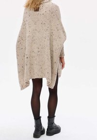 Beige stickad poncho med strukturerat mönster, öppna sidor och hög krage. Bärs över svarta leggings och kraftiga svarta stövlar.