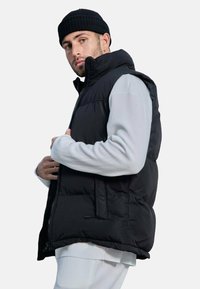 Gilet rembourré noir avec un col haut et une fermeture éclair. Fabriqué en matériau doux et durable, avec des poches latérales et une coupe décontractée.
