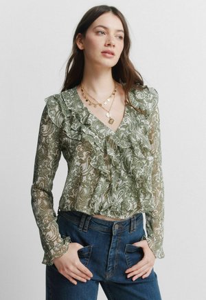 Young woman with long brown hair wearing a green paisley ruffled blouse and blue jeans, hands in pockets, looking to the side.