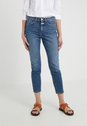 Vrouw draagt mid-rise blauwe cropped skinny jeans, een wit overhemd met opgerolde mouwen en bruine leren sandalen met open tenen.