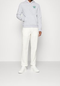 Persona che indossa un hoodie grigio chiaro con il testo "California League", pantaloni bianchi e sneakers bianche, in piedi contro uno sfondo neutro.