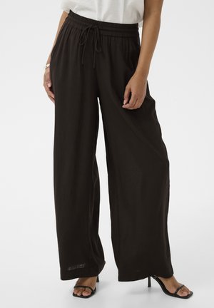 Kvinde iført løstsiddende sorte bukser med snøre og sorte åbne sandaler med hæl, stående mod en ensfarvet baggrund.