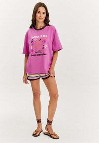 Růžové grafické tričko s textem "Žijící ve svém měkkém chaosu", zkombinované s barevně pletenými šortkami a hnědými sandály na řemínky.