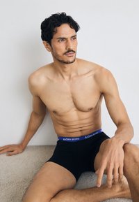 Fekete boxeralsó kék derékgumival, rajta a "BJÖRN BORG" felirattal. A modellen atlétikus testalkat látható, amely egy semleges háttérrel szemben ül.