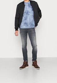Hombre con chaqueta bomber negra, sudadera azul con efecto de lavado, jeans grises desgastados y botas de cuero marrón de pie sobre un suelo gris.