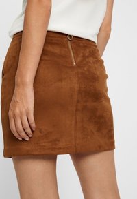 Jupe en daim marron avec une silhouette ajustée, présentant un détail de fermeture éclair sur le côté et une texture lisse, atteignant la longueur mi-cuisse.