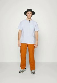 Ανοιχτό μπλε κοντομάνικο πουκάμισο, πορτοκαλί cargo παντελόνι, μαύρο καπέλο bucket και γκρι αθλητικά παπούτσια σε όρθιο ανδρικό μοντέλο με ουδέτερο φόντο.