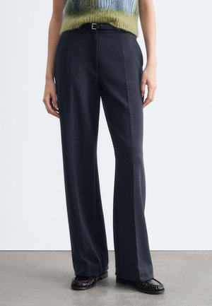 Personne portant un pantalon sombre taille haute à jambes larges, une ceinture noire, des mocassins noirs et un pull sans manches vert et bleu duveteux.