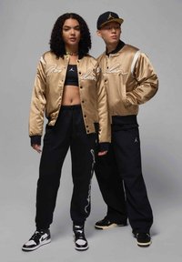 Giubbotti bomber in satin oro con dettagli neri e ricamo "Jordan", abbinati a pantaloni sportivi neri; modello che indossa sneakers.