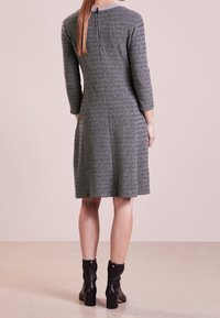 Donna con capelli lunghi in un vestito grigio textured al ginocchio con maniche 3/4 e stivaletti neri con tacco, in piedi di spalle.
