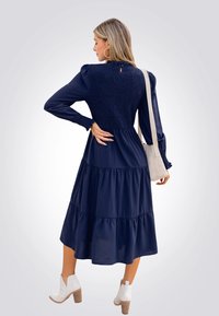 Vestido azul marino con mangas largas, falda en capas y detalles fruncidos en la parte trasera; combinado con un bolso claro y botas blancas hasta el tobillo.