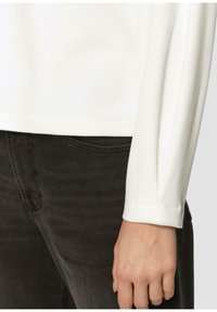 Witte top met lange mouwen en een gladde textuur, voorzien van een eenvoudig ontwerp en een manchet bij de pols, in combinatie met donkergrijze jeans.