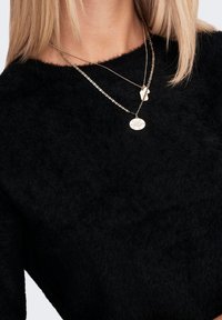 Black fuzzy sweater with a round neck, layered with delicate gold chains featuring pendants, showcasing a textured fabric and metallic accents.