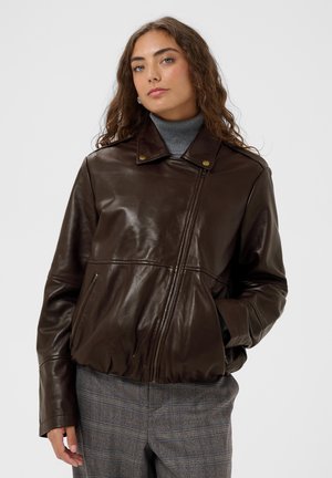 Vrouw met krullend haar die een donkerbruine leren jas draagt over een grijze coltrui en grijze geruite broek, staand tegen een effen achtergrond.