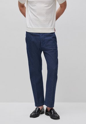 Mann trägt marineblaue, maßgeschneiderte Hose, schwarze Leder-Loafer und ein weißes kurzärmeliges strukturiertes Hemd, steht mit den Händen hinter dem Rücken.