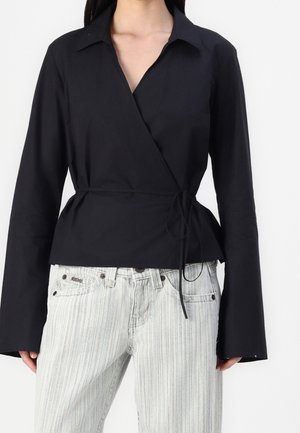 Blouse noire cache-cœur à manches longues, coupe ajustée, avec taille nouée. Associée à un jean en denim clair à rayures, avec boutons et poches décoratives.