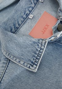 Denimový límec světle modré košile, na které je umístěna bledě růžová etiketa s nápisem "JJXX" a kovové zapínání, s viditelným prošíváním.