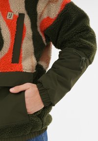 Sudadera de forro polar con un patrón de camuflaje en rojo, beige y verde; cuenta con un bolsillo con cremallera, puños elásticos y un acabado suave y texturizado.