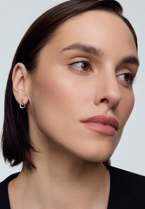 Srebrne kolczyki w kształcie łzy z małymi, przezroczystymi kamieniami, z gładką, wypolerowaną powierzchnią i misternymi detalami. Modelka ma na sobie minimalny makijaż.