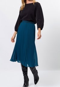 Gonna plissettata turchese realizzata in tessuto leggero, abbinata a una maglietta nera a maniche lunghe e stivali neri fino al ginocchio. Vita accentuata.