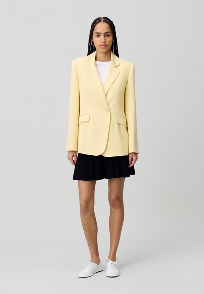 Blazer giallo con un solo bottone, due tasche frontali, abbinato a una gonna plissettata nera e scarpe bianche, indossato da un modello su uno sfondo neutro.