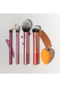 Quatre pinceaux de maquillage de tailles variées et une éponge à estomper orange avec des échantillons de fond de teint et de rouge à lèvres sur une surface claire.