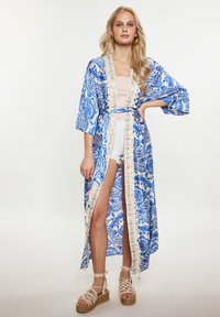 Kimono blu con motivo paisley, maniche larghe, cintura e orlo ricamato. Indossato sopra un top leggero e shorts in denim. Sandali con zeppa intrecciati.