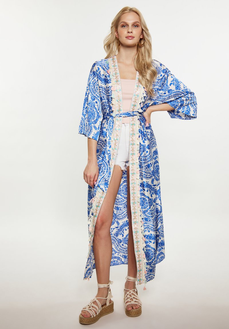 Kimono blu con motivo paisley, maniche larghe, cintura e orlo ricamato. Indossato sopra un top leggero e shorts in denim. Sandali con zeppa intrecciati.