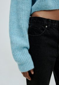 Hellblau gestrickter Pullover mit gerippten Bündchen, kombiniert mit schwarzen Denim-Jeans, die über eine vordere Tasche und einen Metallschlussknopf verfügen.