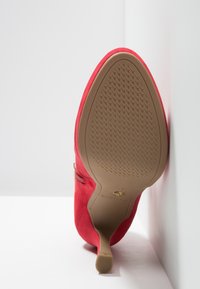 Chaussure à talon haut en daim rouge positionnée semelle vers le haut contre des murs blancs en angle, montrant une semelle marron texturée et un talon fin.